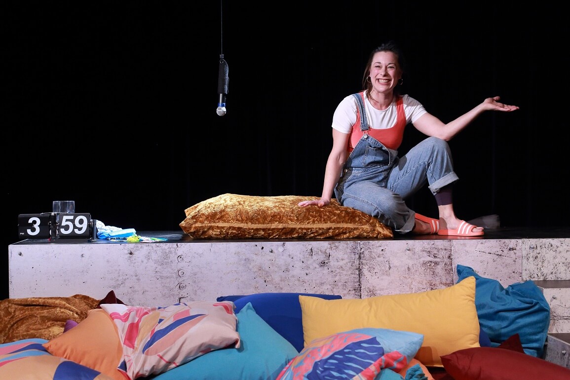 Eine Frau sitzt auf einem Podest, vor ihr auf dem Boden befinden sich viele bunte Kissen. Auf dem Podest ist eine Uhr zu sehen und von der Decke hängt ein Mikrofon herab