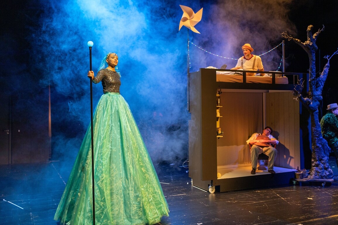 Eine Frau in einem langen blauen Rock und einem langen Zauberstab in der Hand steht vor einem Bühnenbildmodul. Im unteren Teil schläft ein Mann auf einem Sessel, darüber sitzt ein Mann auf einem Bett und schaut zu der Frau