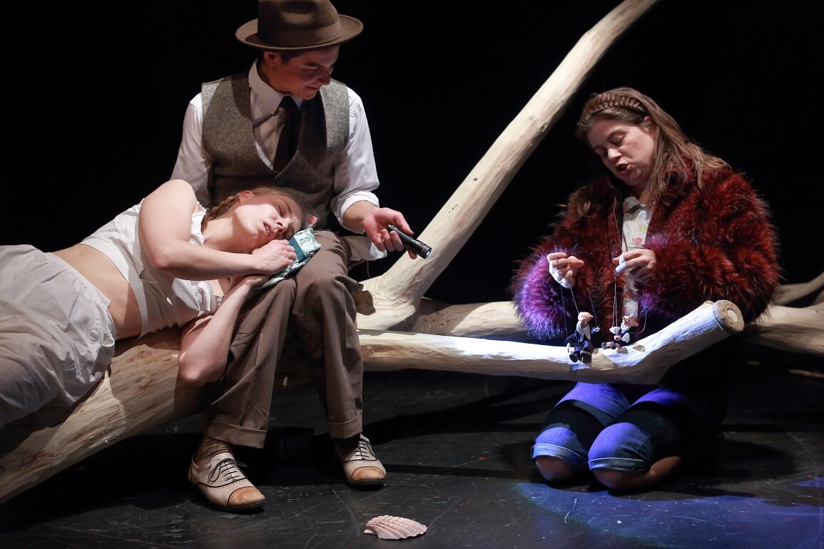 Ein Mädchen liegt schlafend auf einem Baumstamm und mit dem Kopf im Schoß eines Mannes, der eine Taschenlampe in der Hand hält und auf zwei kleine Marionetten leuchtet, die von einem zweiten Mädchen bespielt werden