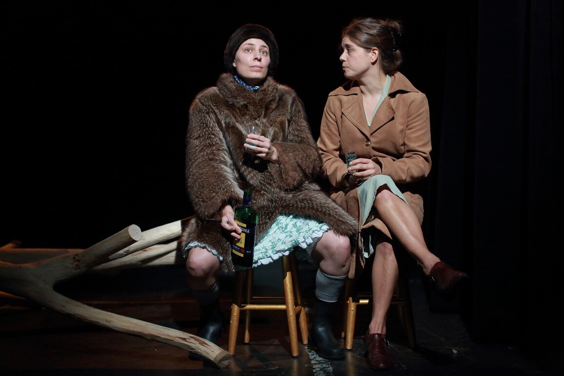 Zwei Frauen sitzen auf Stühlen nebeneinander. Die links sitzende Frau schaut in die Ferne, während die andere sie anschaut