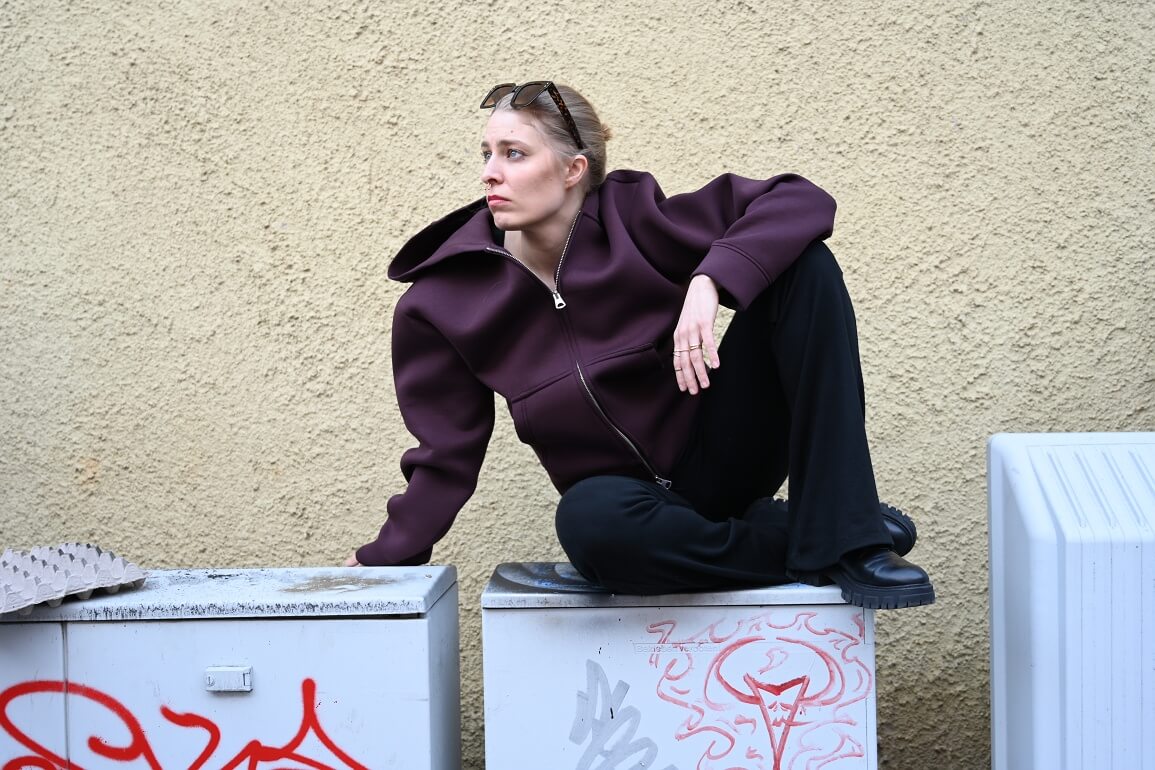 © Maria Clarissa Wolf Eine Jugendliche in lockerer schwarzer Hose und weitem Kapuzenpulli sitzt mit angezogenen Knien auf mit Graffiti besprühten Stromkästen und schaut zur Seite