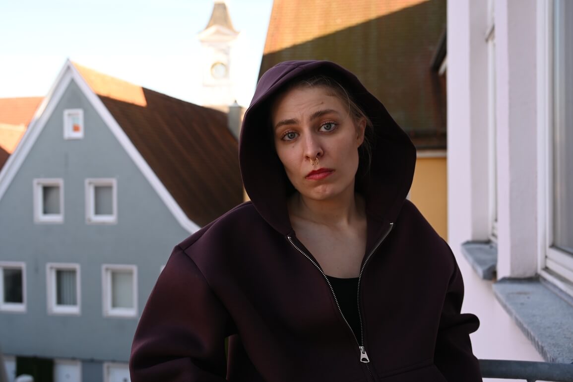 © Maria Clarissa Wolf Eine Jugendliche schaut mit skeptischem Blick in die Kamera, sie trägt einen weiten Kapuzenpulli mit der Kapuze über dem Kopf und hat ein Nasenpiercing
