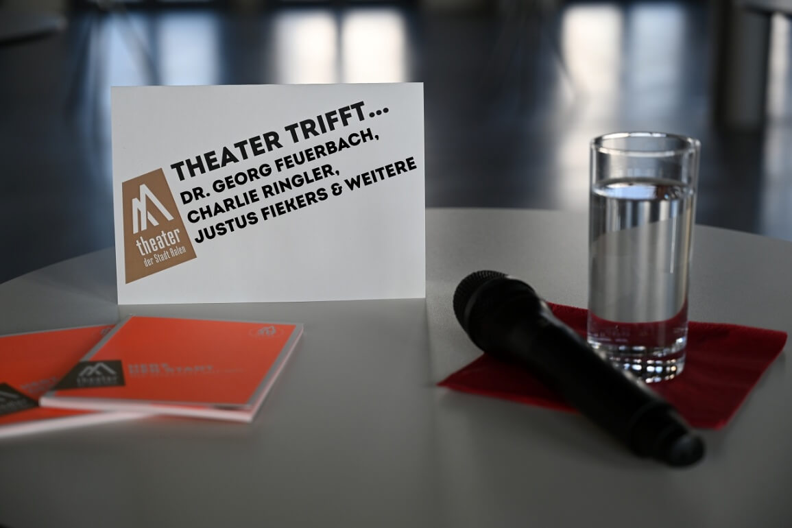 © Robin Kucher Auf einem Tisch liegt ein Mikrofon neben einem Wasserglas und Spielzeitheften des Theaters. Auf einem Schild ist das Logo des Theaters sowie "Theater trifft... Dr. Georg Feuerbach, Charlie Ringler, Justus Fiekers & weitere" zu lesen