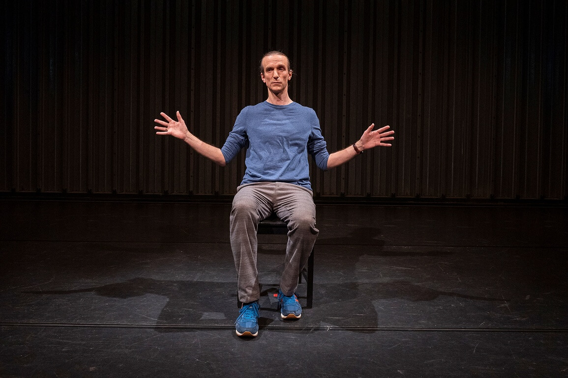 Ein Mann in blauem Pulli sitzt auf einem Stuhl, die Arme seitlich ausgestreckt, die Finger gespreizt