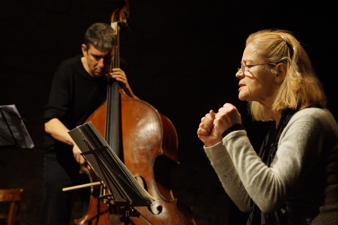 Eine Frau sitzt vor einem Notenständer und liest davon ab, im Hintergrund spielt ein Mann Kontrabass