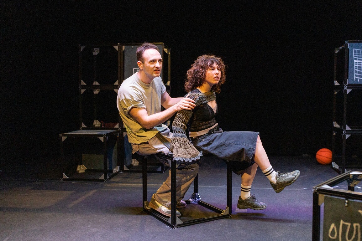 Eine männlich und eine weiblich gelesene Person sitzen seitlich auf einem Metallkubus. Die männlich gelesene Person hat ihre Hände auf den Schultern der weiblich gelesenen Person, die ihre Beine angehoben hat und beide schauen in die Ferne