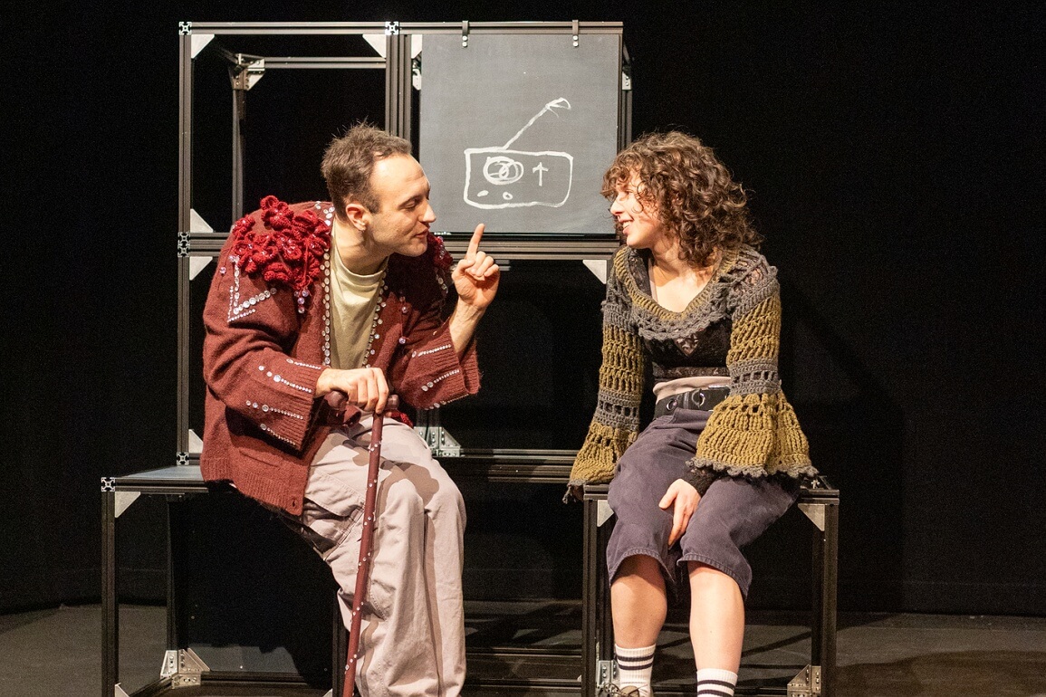 Eine männlich und eine weiblich gelesene Person sitzen auf zwei Metallklötzen. Aus diesen ist auch ein Turm hinter ihnen gebaut, auf einem Schild ist ein Radio gemalt. Die männlich gelesene Person ist auf einen Stock gestützt und hebt den Zeigefinger