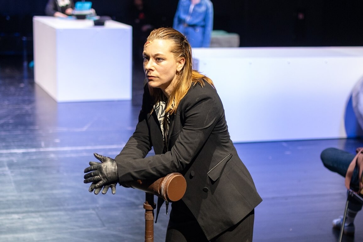 Eine Frau in einem schwarzen Anzug mit schwarzen Lederhandschuhen, zurückgegelten Haaren und einer Wunde am Kopf stützt sich mit den Unterarmen auf einem überdimensional großen Richterhammer ab