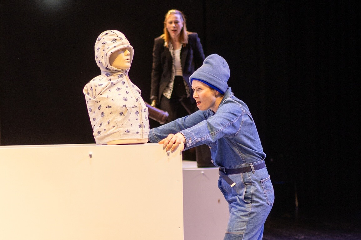 Auf einem weißen Podest steht ein Puppentorso, auf das Podest stützt sich eine Frau in einem blauen Overall ab und schaut die Puppe an, im Hintergrund ist ebenfalls auf einem Podest eine Frau in einem schwarzen Anzug und Richthammer zu sehen