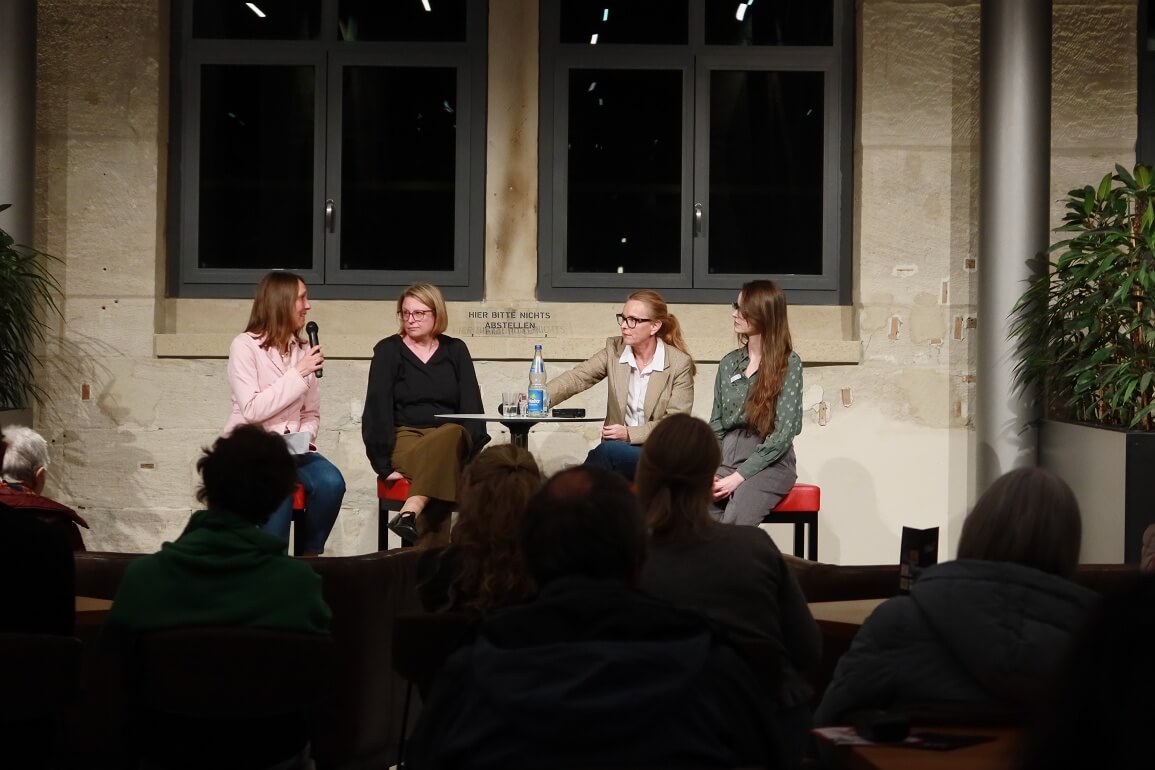 © Maria Clarissa Wolf Vier Personen sitzen auf Barhockern um einen Stehtisch herum, die Person ganz links spricht in ein Mikrofon, die anderen hören ihr aufmerksam zu. Im vorderen Bereich sieht man mehrere Menschen von hinten, die zu den vier Personen blicken