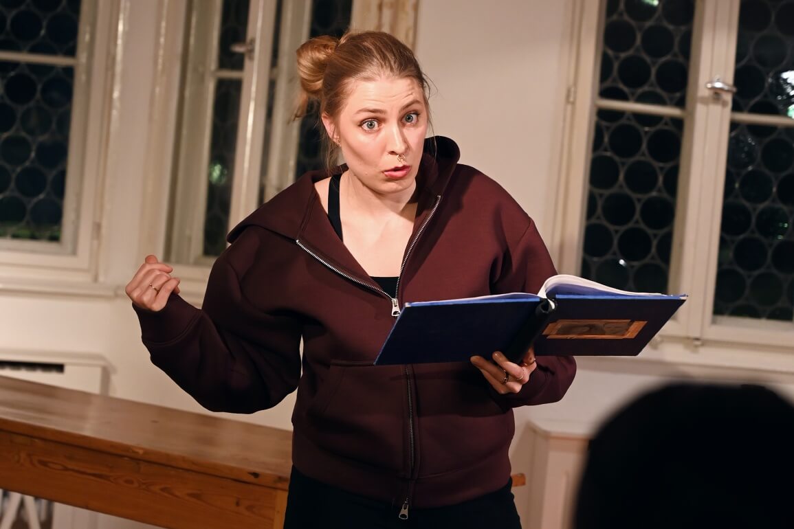 © Maria Clarissa Wolf Eine Frau hält ein aufgeschlagenes blaues Buch in der linken Hand und schaut mit eindringlichem Blick auf eine Person vor ihr, während sie mit der rechten Hand hinter sich zeigt