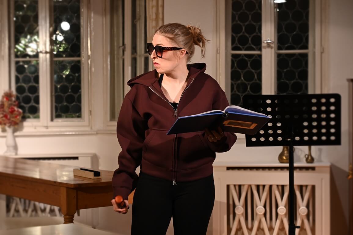 © Maria Clarissa Wolf Eine Sonnenbrille tragende Frau hält ein aufgeschlagenes blaues Buch und ein Mikrofon in der Hand und schaut zur Seite