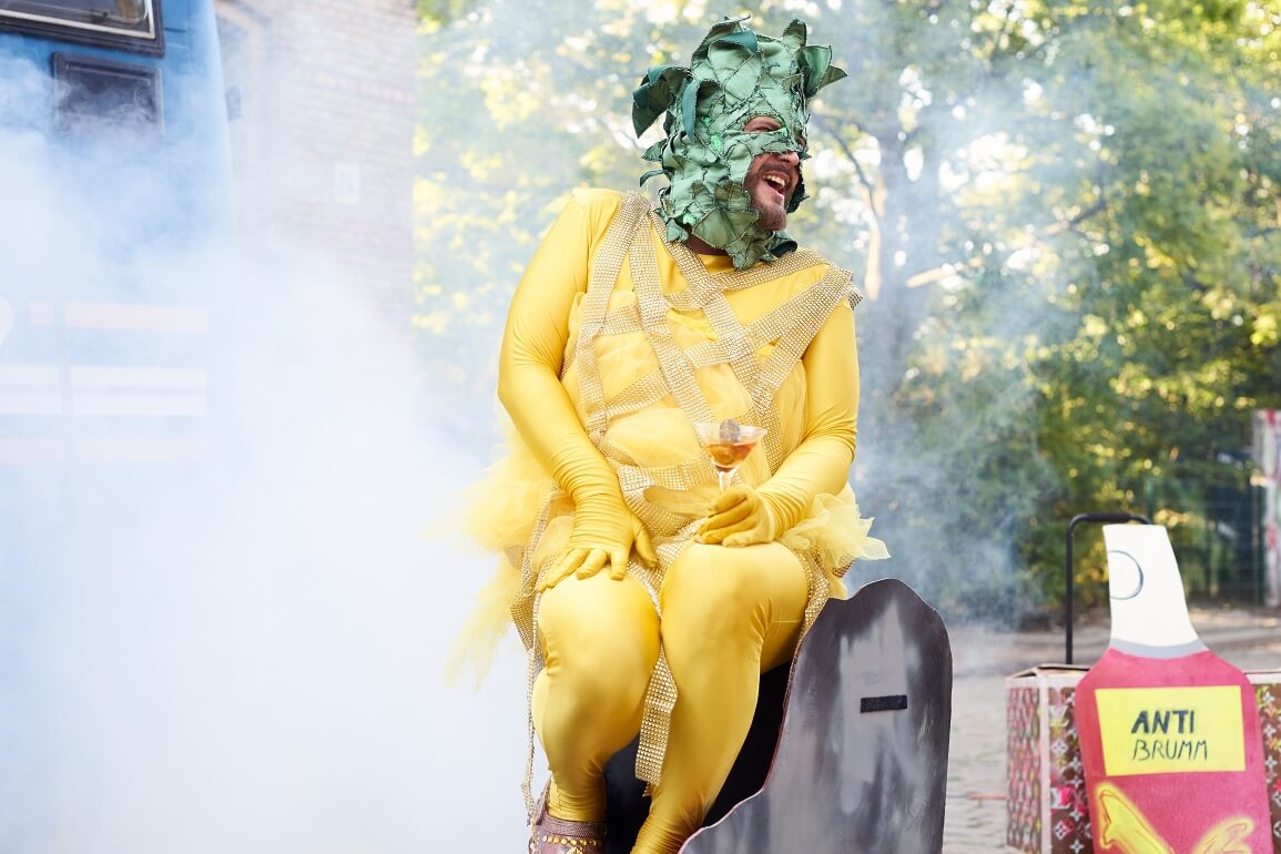 eine Person in einem gelben Ganzkörperanzug und einer grünen Maske sitzt auf einem Stuhl, hinter ihm sind Bäume und Nebel zu sehen