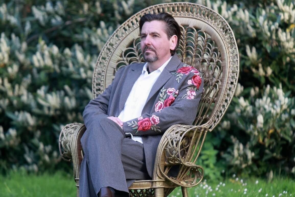 Ein Mann in einem mit Blumen bestickten Anzug sitzt auf einem großen, goldenen Stuhl auf einer Wiese, im Hintergrund blüht ein weißer Fliederbusch