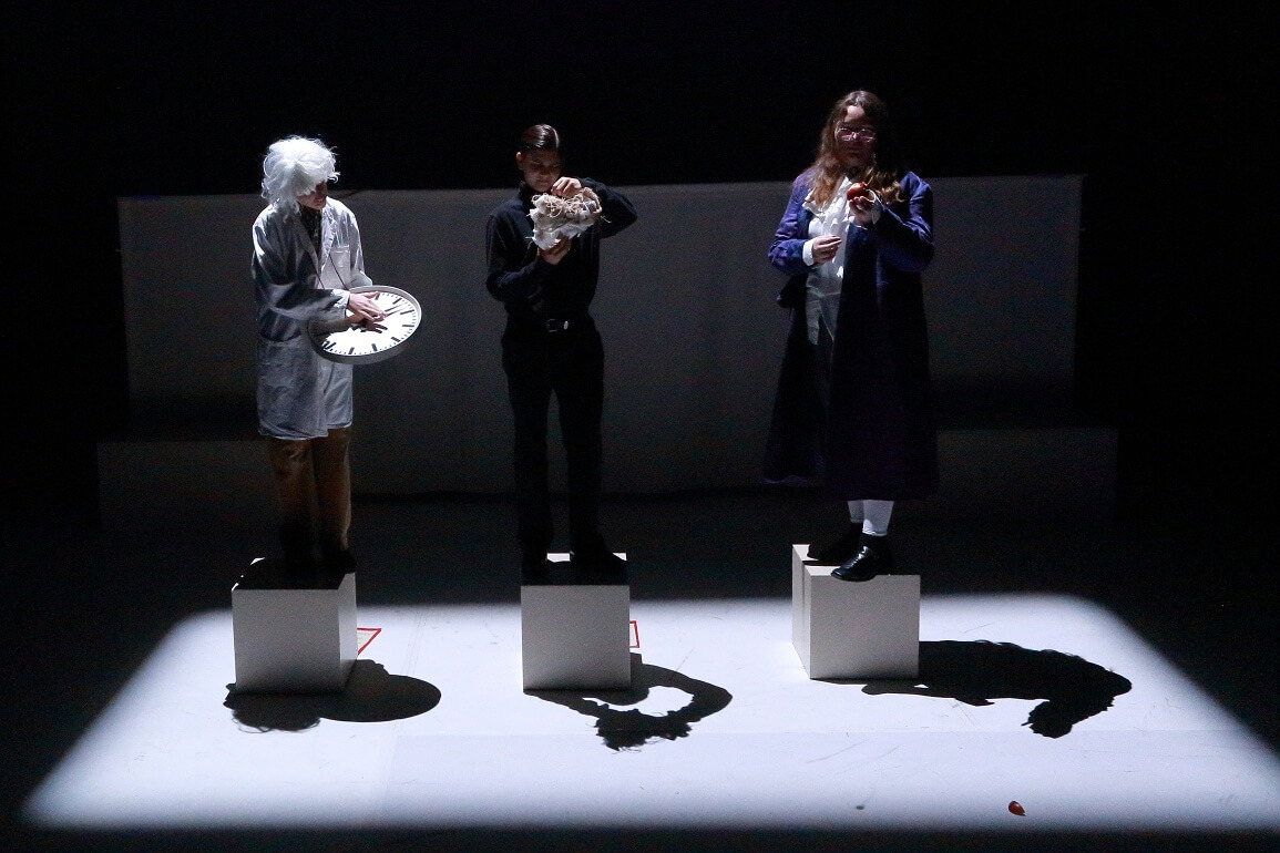 © Peter Schlipf Drei Personen stehen, jeweils mit einer Uhr, einer Maske und einem Apfel in der Hand, auf drei Hockern und werden mit grellem, weißem Licht angeleuchtet