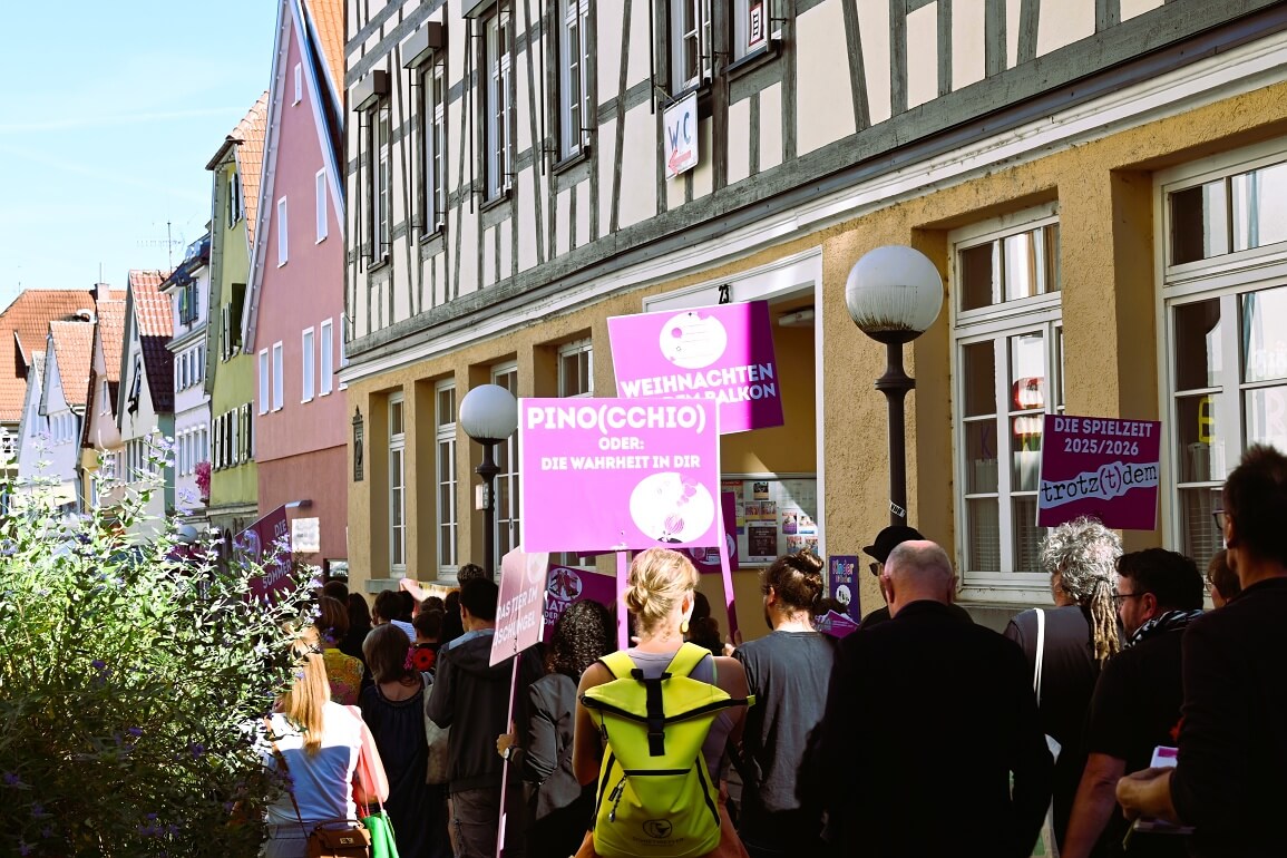 Menschen laufen mit Schildern, auf denen die Titel von Theaterstücken zu lesen sind, durch eine von Fachwerkhäusern geprägte Altstadt