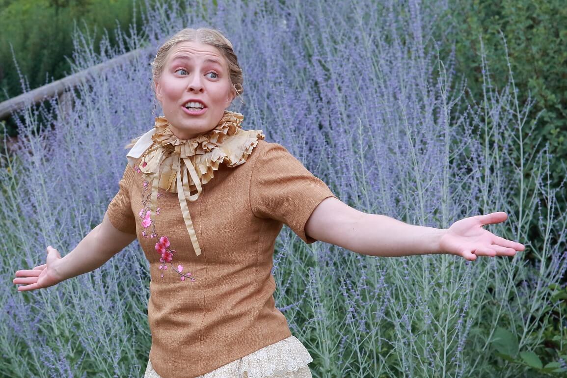 Eine Frau in einem goldenen Kostüm breitet die Arme zu beiden Seiten aus, im Hintergrund sind blau blühende Blumen zu sehen